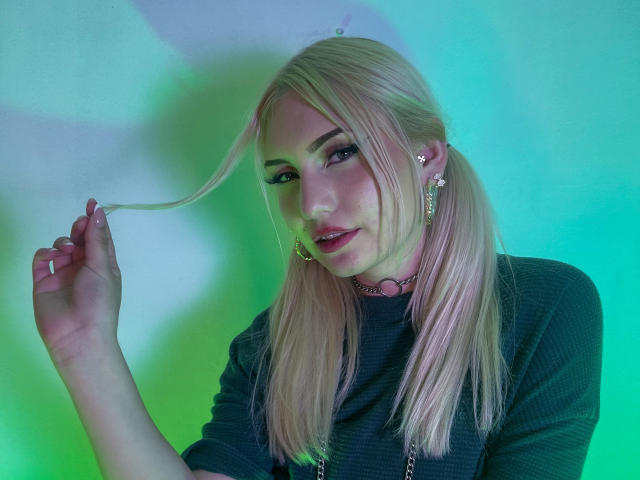 Fotografia de MariyLu, loira, com cabelo liso, segurando uma mecha, com luzes verdes e azuis iluminando seu rosto. Usando uma blusa preta, um colar rosa e brincos. Expressão sexy, confiante e ligeiramente sedutora..