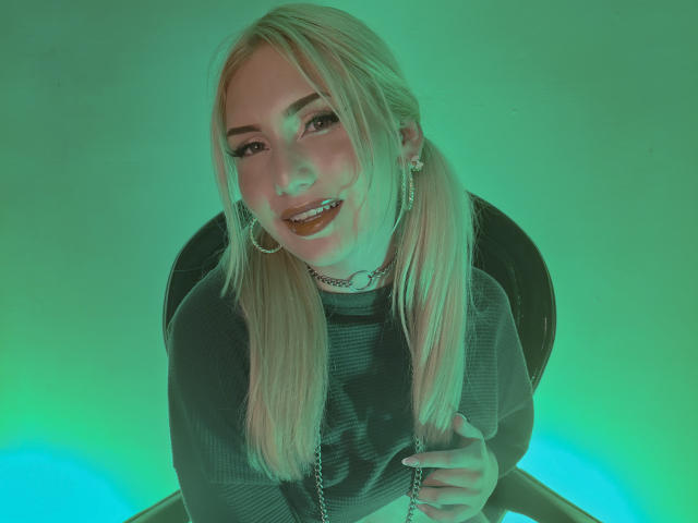 Foto van MariyLu, een blonde vrouw met lang haar, die een zwarte trui draagt en op een stoel zit, met een groene-blauwe gloed. licht van onderen, lachend met een hand op haar borst, sensuele uitstraling.