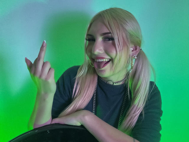 MariyLu with platinum blonde pigtails, black shirt, and choker, flashing a middle finger with a wide, toothy grin. Neon green and blue light background, creating a vibrant, edgy vibe.