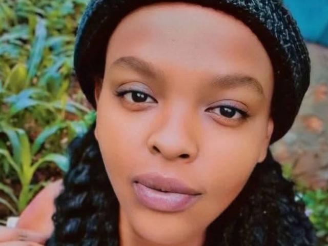 Close-up of Tinna-hot, dark-skinned with braided hair, wearing a black beanie, staring directly at the camera with neutral expression, full lips, slight shadow under eyes, greenery in background. Fucking sexy.