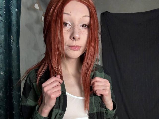 Close-up photo of HannaCool, a pale-skinned woman with red hair, freckles, and a nose ring, gripping her green shirt while looking at the camera. Her expression is slightly seductive.
