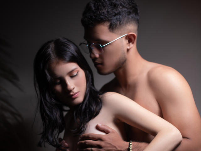 Fotografía de EimyAndMaxi, un hombre sin camisa con cabello negro corto y gafas, agarrando a una mujer sin top y con cabello negro largo. cabello, presionando su pene contra su pecho, iluminación oscura, enfoque intenso..