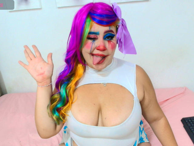 Foto van GraceYuly, een mollige vrouw met fel gekleurd regenbooghaar, een groot decolleté in een witte mouwloze top, die zwaait. Tong uitgestoken, blauwe en roze gezichtsbeschildering, kleurrijke rok met patroon..