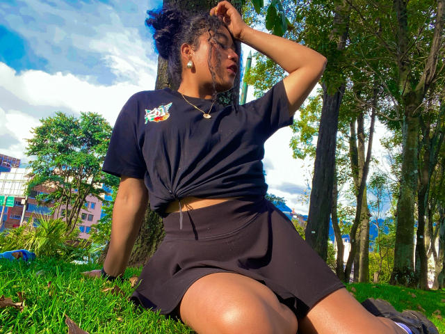 SophieSonyas, une femme à la peau foncée avec des cheveux bouclés, est assise sur l'herbe, une main dans ses cheveux, portant un t-shirt noir noué à la taille, noir shorts. La lumière du soleil met en valeur ses courbes, avec des arbres en arrière-plan. Incroyablement sexy..