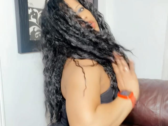 Cincia69 met lang, krullend zwart haar dat haar gezicht bedekt, draagt een zwarte top zonder bandjes en raakt haar borst met één hand aan. rode polsband, bruine leren stoel, witte achtergrond, seksuele spanning.