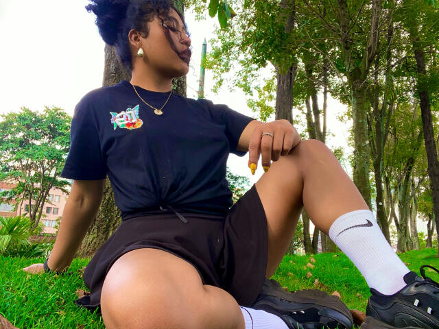 SophieSonyas sitting on grass with legs spread, wearing black t-shirt, black shorts, white socks, and black shoes. Her hand rests on knee, cock slightly visible, forest background. Fucking sexy.