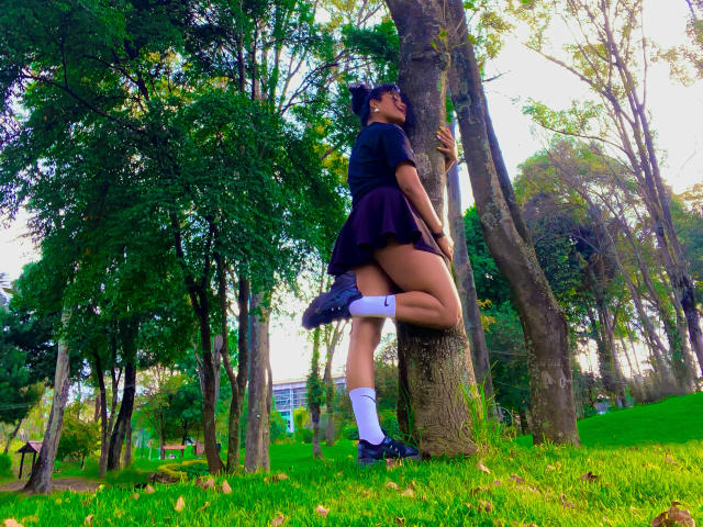 SophieSonyas, standing with legs wrapped around a tree trunk, wearing a black dress, white socks, and black shoes, her bare thighs exposed, green park background, trees, sunlight filtering through leaves.