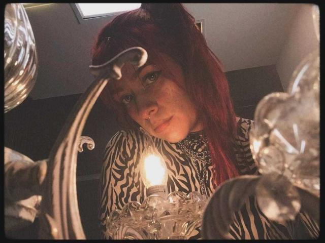 Close-up van LolyYunno met rood haar, een shirt met een zebrapatroon en een intense blik, die naar beneden kijkt naar een brandende kaars, omringd door versierde glazen armaturen. Schaduwen versterken de intieme, prikkelende sfeer..