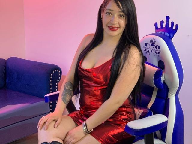 Alanna Scott sitzt auf einem weißen Gaming-Stuhl mit einer blauen Rückenlehne und trägt ein eng anliegendes, glänzendes rotes Kleid, das tiefe Ausschnitte zeigt. Schwarzes Haar, roter Lippenstift, Tätowierung am linken Arm, lächelnd. Rosa und blaue Beleuchtung..