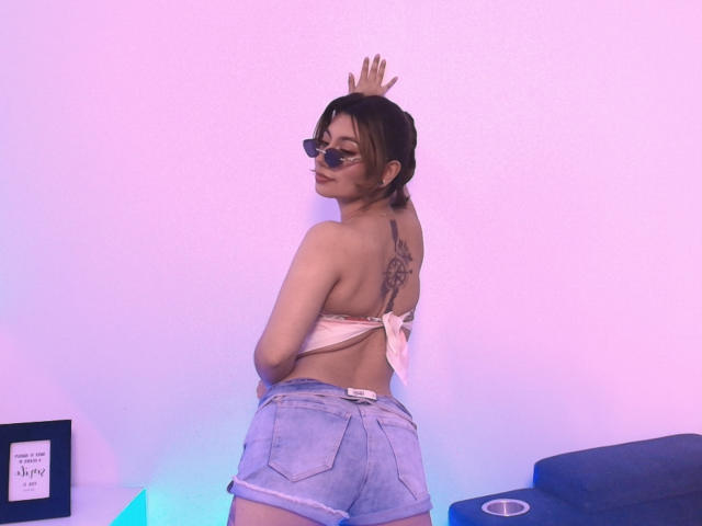 Selene Leon, de espaldas a la cámara, vestida con un top de bikini rosa y pantalones cortos vaqueros azul claro, de pie frente a un fondo blanco. pared. Tatuaje en la parte superior de su espalda, mirando por encima del hombro, pose seductora..