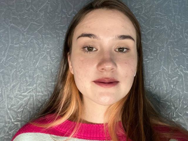 Fotografia de um close-up de VertieTllo com cabelos castanhos longos, vestindo um suéter listrado em vermelho e branco, com um fundo texturizado. Fundo azul-acinzentado. Ela olha diretamente para a câmera, com um leve sorriso. Sua pele é clara e ela tem sardas visíveis. A iluminação realça o rosto dela e cria uma leve sombra no lado esquerdo. Sua expressão é neutra, mas ligeiramente convidativa. O foco está nítido e as cores são vibrantes. A imagem tem um leve efeito de "falha" na textura do fundo..