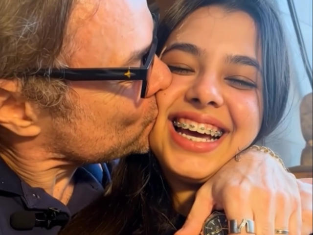 Close-up of ViktorAndPenny with a man kissing her cheek. She's smiling with braces, brown hair, and a ring on her right hand. He wears black sunglasses and a dark shirt.
