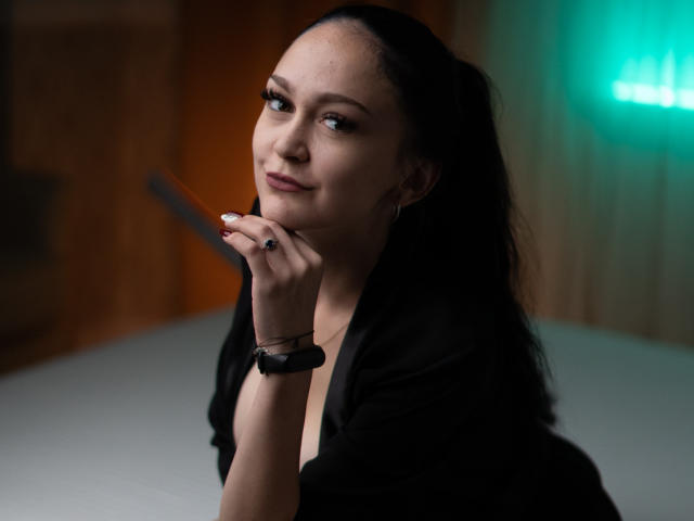 Fotografía de BustyBabeHot con cabello oscuro y ondulado, con una ligera sonrisa, mano cerca de la barbilla, vistiendo una blusa negra, con una pulsera en la mano izquierda. Muñeca, tenuemente iluminada con luz verde en el fondo. Tensión sexual evidente..