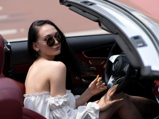 BustyBabeHot with long black hair, wearing off-shoulder white top, round sunglasses, yellow nails, sitting in car, hand on steering wheel, car door open, sexy pose, sunlight highlights her.