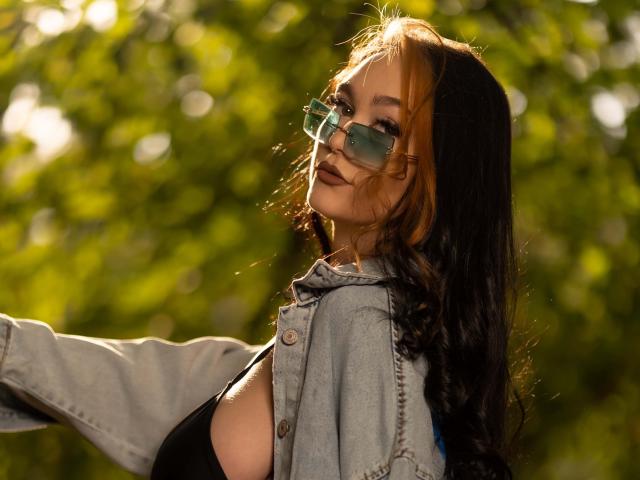 Fotografia de BustyBabeHot com cabelos castanhos longos, usando óculos de sol estilo aviador e um casaco jeans azul claro, com luz solar. Iluminando seu rosto, mão estendida para frente, fundo com folhagem verde, leve sombra no peito..