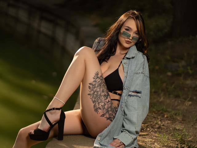 BustyBabeHot sits on the ground, legs spread, wearing a black bra, denim jacket, and black high heels. She has large floral tattoos on her thigh and wears sunglasses. Fucking sexy.