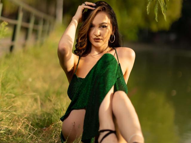 Eine wohlgeformte Frau sitzt auf dem Gras, die Beine gespreizt, die Hand im braunen Haar, trägt ein grünes Kleid, das Sonnenlicht erzeugt Schatten, ihr Blick ist verführerisch, sie trägt einen Reifen. Ohrringe, natürliches Licht, unscharfer Hintergrund, Außenaufnahme, sexuelle Spannung, Sommerstimmung..