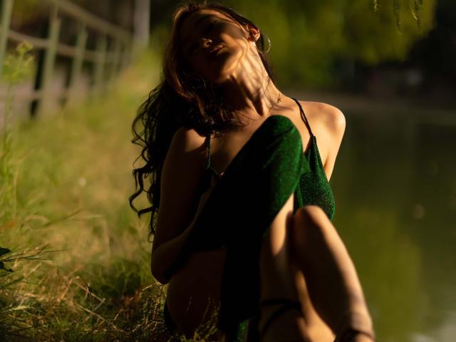 Fotografia de BustyBabeHot, sentada em uma margem gramada, com a cabeça inclinada para trás, cabelos longos e escuros, vestido verde, com a luz do sol incidindo. sombras, pernas cruzadas, forte tensão sexual, fundo desfocado, vegetação, luz natural.