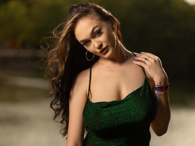 Foto van BustyBabeHot met lang, golvend bruin haar, gekleed in een groene, glinsterende jurk met spaghettibandjes. Ze kijkt verleidelijk in de camera, met haar hand op haar borst, terwijl het zonlicht haar rondingen accentueert..