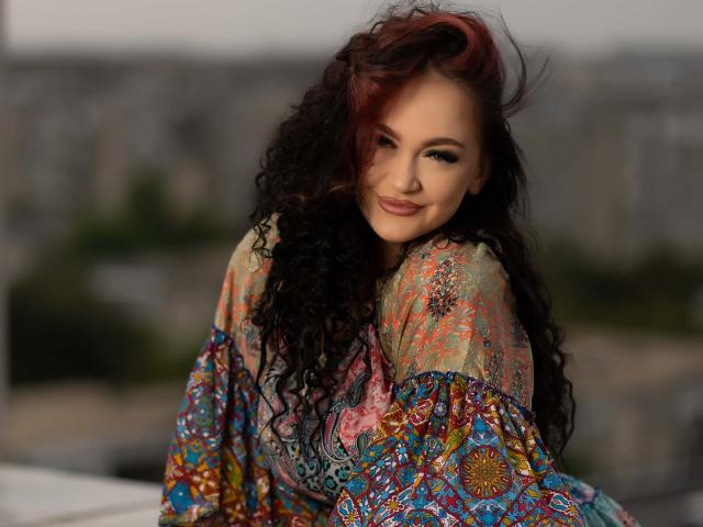 BustyBabeHot with long, curly black hair, wearing a colorful, patterned shawl, looking back with a seductive smile. Blurred cityscape in the background, bright lighting, sexy vibe.