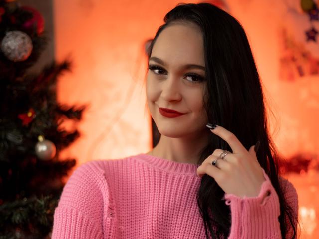 Eine üppige Frau mit langen, schwarzen Haaren, rotem Lippenstift und einem pinken Strickpullover, die verführerisch lächelt, die Hand in der Nähe des Gesichts, unscharf. Weihnachtsbaum und leuchtender, oranger Hintergrund. Verdammt heiß, verführerische Atmosphäre..