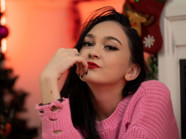 Fotografia de BustyBabeHot com cabelos longos e pretos, batom vermelho e um suéter rosa de tricô, tocando seus lábios com expressão sedutora, brilho vermelho ao fundo, decoração festiva..