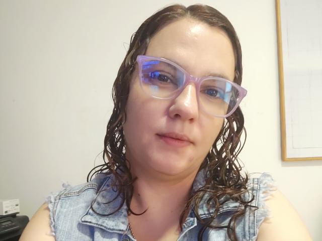 Gros plan d'Amelia Wilson, avec les cheveux bruns mouillés, portant des lunettes transparentes et un gilet en jean. Son expression est neutre, avec une légère tension sexuelle indiquée par ses lèvres détendues et légèrement entrouvertes..