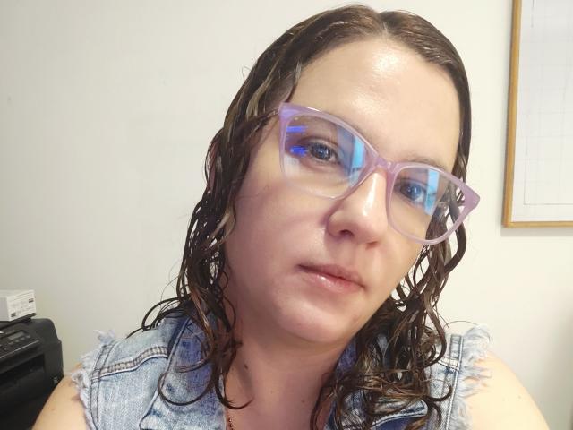 Primo piano di Amelia Wilson con i capelli castano scuro e bagnati, che indossa occhiali trasparenti e una camicia a quadri grigia. La sua espressione è leggermente seducente, con un accenno di tensione sessuale. Illuminazione intensa.