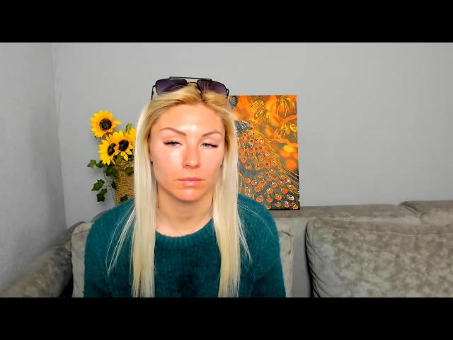 Photograph of FortyLeride with long blonde hair, wearing a black top, sitting on a gray couch. Sunflowers in a vase and a colorful abstract painting in the background. Fucking relaxed expression.