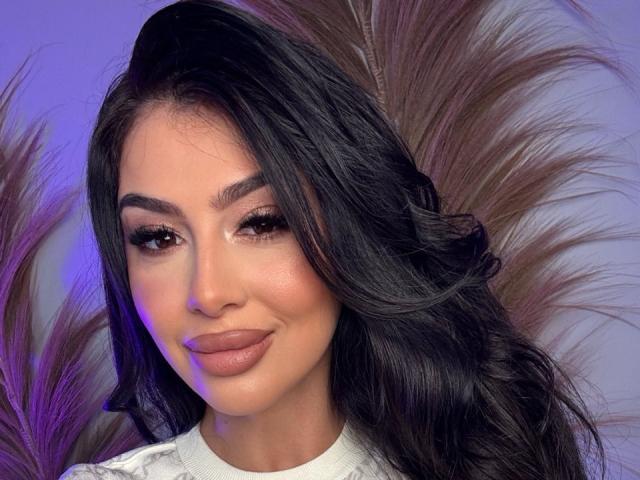 Fotorealistisches Porträt von MissMeganMoon mit langen, welligen schwarzen Haaren, vollen Lippen und dunklen Augenbrauen. Sie blickt verführerisch in die Kamera, mit einem leichten Lächeln, umgeben von zarten, violetten Akzenten..