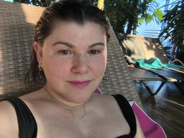 Primer plano de SportyLove con cabello castaño oscuro y húmedo, piel clara y lápiz labial rosa, sentada en una silla de mimbre, con una correa negra. vista frontal, collar dorado, ligera sonrisa, luz del sol filtrándose entre los árboles.