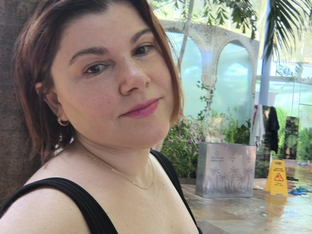 Photograph of SportyLove, a plus-sized woman with fair skin and brown hair, wearing a black tank top, gold necklace, and earrings, with a slight smirk, surrounded by outdoor plants and a wet floor sign.