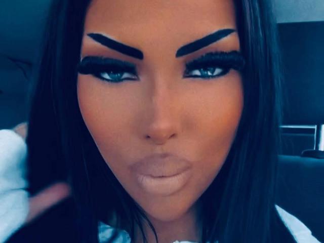 Gros plan sur LuxuryDoll69, une femme aux cheveux foncés, avec des sourcils noirs épais, un trait d'eye-liner prononcé et des yeux bleus. Elle regarde directement l'appareil photo avec une expression boudeuse. Regard concentré, intense.
.