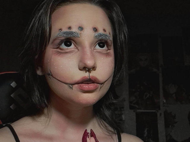 Close-up of KenyattaConsigli with dark hair, pale skin, and blue eyeshadow. She has a nose ring, black lines on her face, and red marks on her chest. Dark, moody lighting.