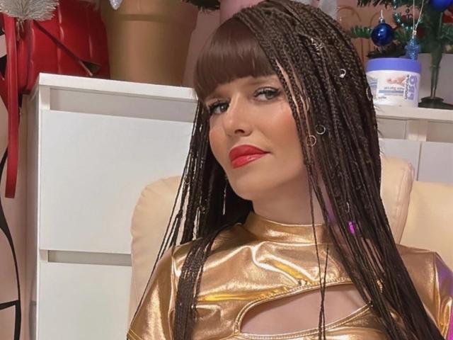 Fotografía de primer plano de CutieOrgasm con cabello largo y trenzado, vistiendo una blusa dorada brillante con detalles de corte en los hombros, de color rojo intenso. labial, y una expresión de confianza. El fondo incluye muebles blancos y tela roja..