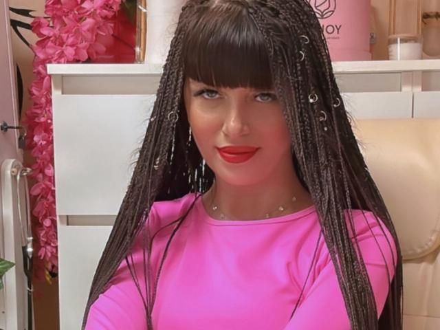 Photograph of CutieOrgasm with long braided hair, wearing a bright pink shirt, red lipstick, and a collar with small studs. She's looking directly at the camera with a seductive smile.
