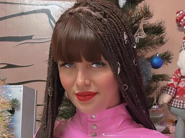 Close-up foto van CutieOrgasm met lange, donkere vlechten, felrode lippenstift en een roze top met knoopjes. De achtergrond toont een versierde kerstboom met blauwe versieringen en een kerstmanpop..