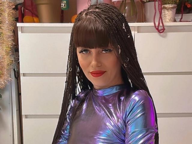 Close-up foto van CutieOrgasm met lang gevlochten haar, die een glanzende blauwe latex top draagt, rode lippenstift en kleine oorringen. oorringen. De achtergrond toont witte planken met diverse objecten. Een verleidelijk en aantrekkelijk beeld..