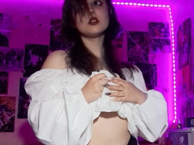 KenyattaConsigli, senza reggiseno e con lunghi capelli neri, indossa una camicia bianca con una spalla scoperta, tenendola aperta, rivelando il seno. seni. Luci viola neon, poster appesi alle pareti, tensione sessuale, rossetto rosso scuro.