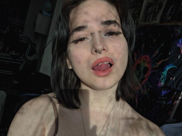 Foto em close-up de KenyattaConsigli, uma mulher de pele clara com cabelo preto, piercings no nariz e piercing no lábio. Ela está esticando a língua com uma expressão sexual e provocante. Padrões de sombra em seu rosto.