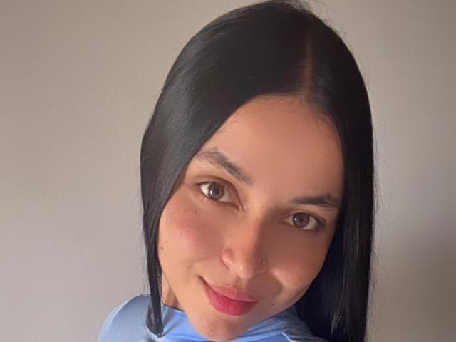 Nahaufnahme von HikariChloe, einer Frau mit langen schwarzen Haaren, braunen Augen und rotem Lippenstift, die leicht lächelt und ein helles blaues Hemd. Ihr Gesicht ist gerötet, mit sichtbaren Sommersprossen auf den Wangen..