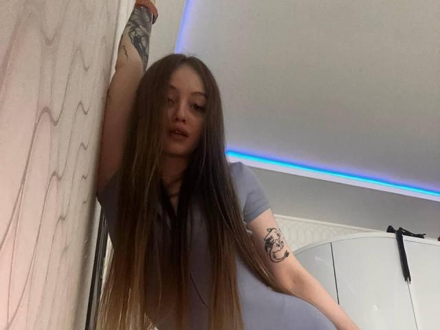 Jacqulyn Burtis, com cabelos castanhos longos e lisos, apoiada em uma parede, com o braço levantado, mostrando uma tatuagem no antebraço. Ela está vestindo uma camisa cinza, com uma luz LED azul ao fundo. Incrivelmente sexy..