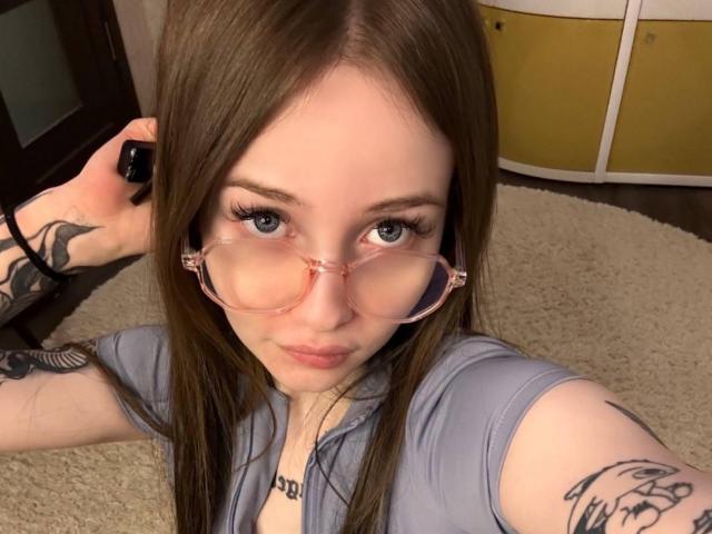 Nahaufnahme-Selfie von JacqulynBurtis, einer jungen Frau mit heller Haut, langen braunen Haaren und rosa Brille, die ein Telefon hält. sichtbare Tätowierungen am Arm, trägt ein hellblaues Hemd, verführerischer Blick.