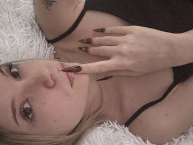 Close-up de ElidiaKrotine deitada em uma superfície branca e fofa. Ela tem cabelo loiro, pele clara e esmalte preto. Sua mão está perto da boca, mostrando uma expressão sedutora. Ela está vestindo uma blusa preta..