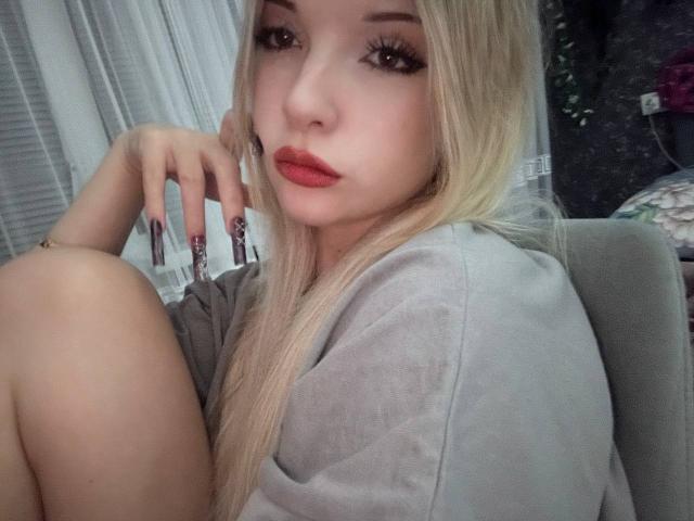 Close-up of ElidiaKrotine, blonde with long hair, red lipstick, heavy eyeliner, and long black nails. She's sitting, legs bent, wearing a grey shirt, looking seductively at the camera.