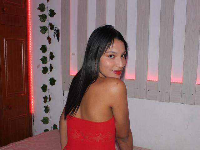 Cloe Boyer, mit dem Rücken zum Betrachter, blickte über ihre Schulter, trug ein rotes Spitzenkleid ohne Träger, hatte schwarzes Haar und trug roten Lippenstift. helle, braune Haut, Wand mit weißen und rosa Streifen, rote LED-Leiste, Blattdekoration.