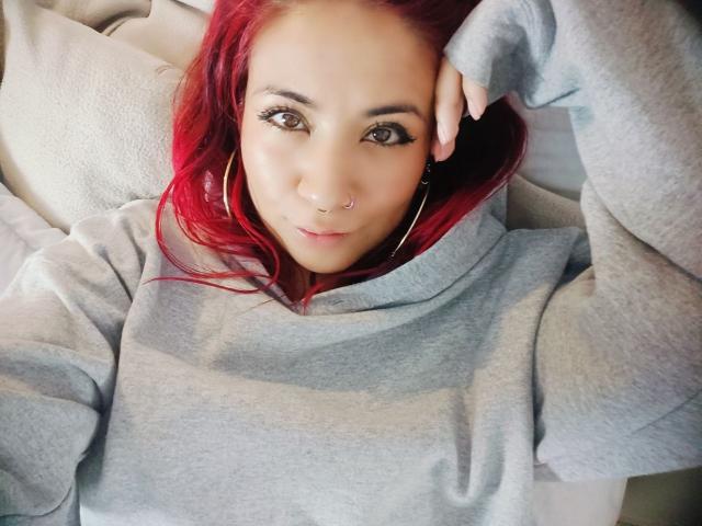 Larissa Miller, uma jovem de cabelos ruivos, está deitada em um sofá branco, vestindo um moletom cinza. Ela tem um piercing no nariz, brincos de argola e olhos castanhos intensos. Incrívelmente atraente. Mão no cabelo..