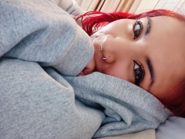Close-up of LarissaMiller with red hair, piercing nose ring, and dark eyebrows, lying on a bed, partially covered by a gray blanket, with a suggestive, relaxed expression. Bright sunlight highlights her face.