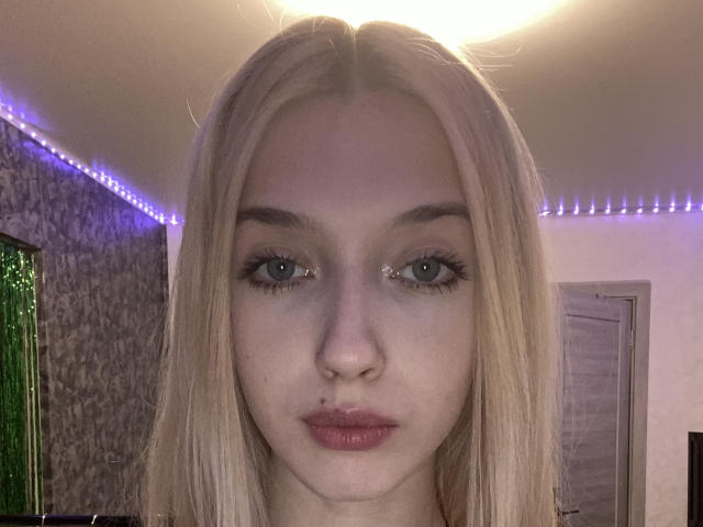 Close-up van MarcellaMcleskey, blond met steil haar, bleke huid, blauwe ogen en volle lippen, die recht in de camera kijkt. camera. De achtergrond bestaat uit paarse LED-strips en een gestructureerde muur. Extreem intense blik.