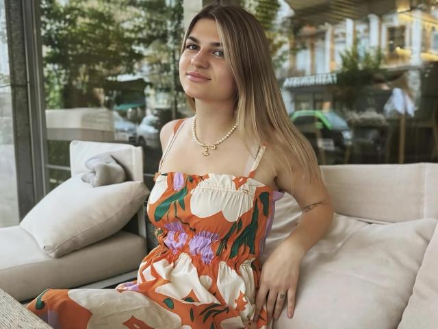 CrystalBlush sitting on a white couch, wearing a colorful floral dress with spaghetti straps, pearl necklace, and a bracelet. She has straight brown hair, light skin, and a subtle smile. Background shows a sunny outdoor cafe.